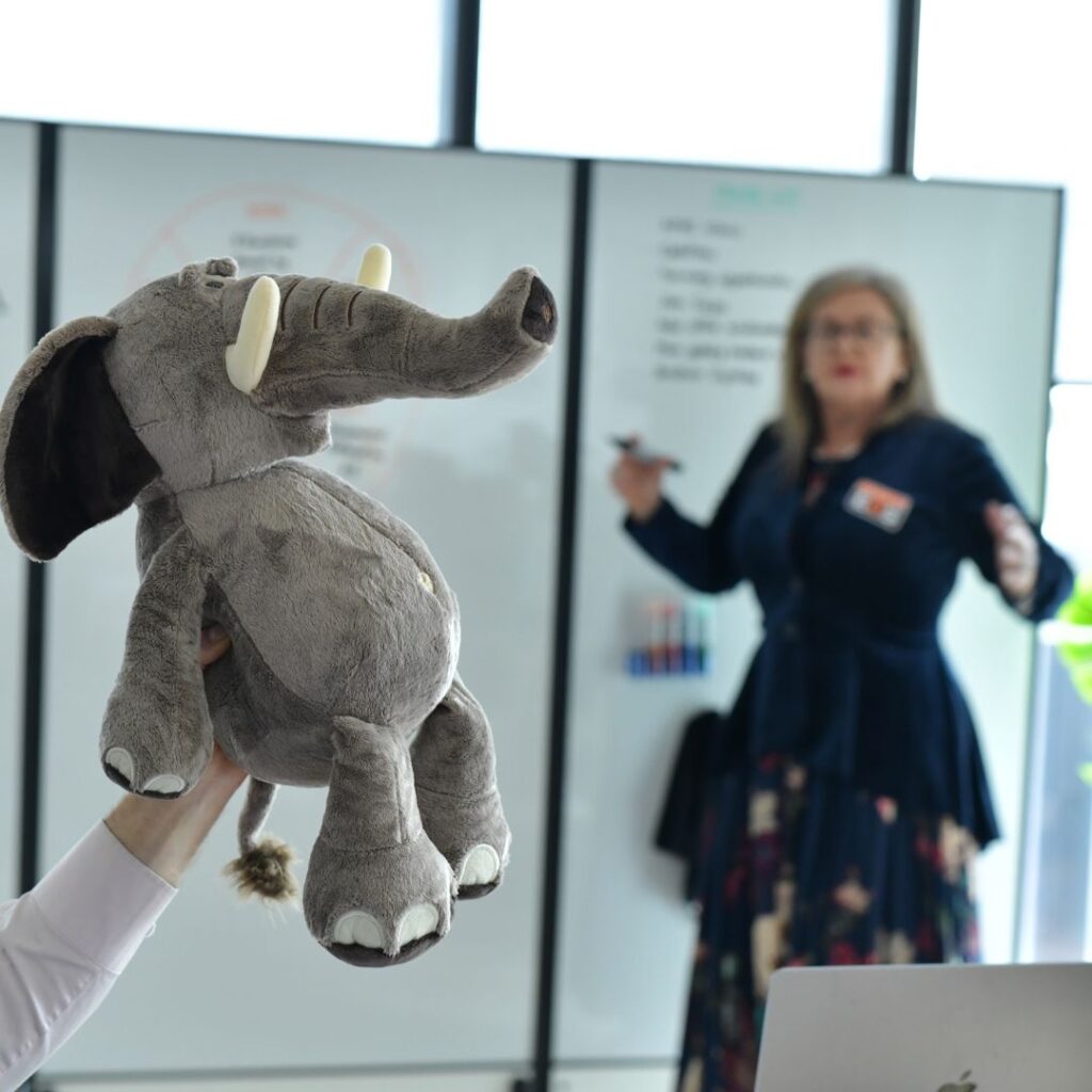 Plush elephant toy in the foreground with a woman writing on a whiteboard in the background.