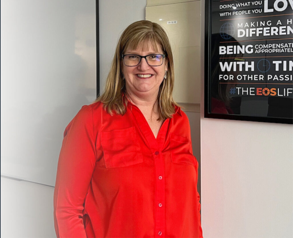 Woman in a red blouse smiling while standing next to a motivational poster in an office setting.