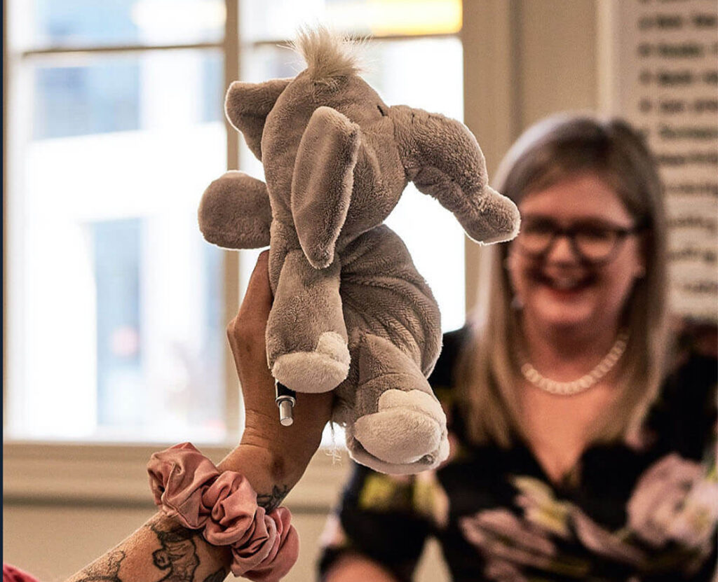 Plush elephant toy held up in the foreground with a smiling woman in the background.